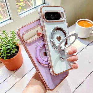 May include: Two smartphone cases with rose gold metallic edges. One case is white with a central magnetic ring, a heart icon, and the word nice printed on the side. The other case is lavender with a matching magnetic ring. Both feature heart patterns.