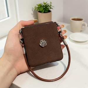 May include: A square, dark brown textured case with a gold-toned metal flower emblem centered on the front. The case features silver-toned metal hardware attachments on the sides and a thin, smooth brown strap connected to the sides.