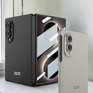 May include: A black foldable smartphone case with a camera cutout and the text FOLD CARE DESIGNED BY GKK, alongside a separate light gray foldable smartphone case featuring the same camera layout and the text FOLD CARE DESIGNED BY GKK.