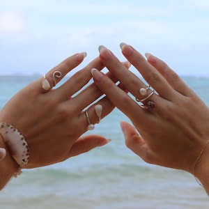 Puede incluir: Una colección de anillos de plata con pequeñas perlas blancas y un diseño en espiral, usados junto a una pulsera adornada con una concha grande y estampada. Las piezas de joyería son metálicas y blancas, con formas orgánicas y texturas lisas.