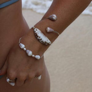 Puede incluir: Un brazalete de plata con dos conchas cónicas pulidas en los extremos, una pulsera de cadena adornada con pequeñas perlas blancas y conchas cónicas, y un anillo con una perla y una concha pequeña, todo en tonos naturales marrones y blancos.
