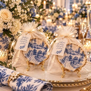 May include: Two decorative scallop shells featuring blue and white landscape illustrations, each displayed on a gold metal stand inside a sheer organza drawstring bag with a white tag printed with the text Thank you for being part of our journey.