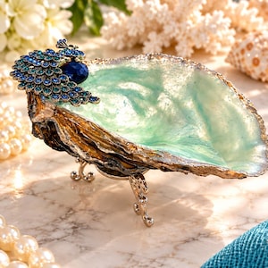 May include: A decorative oyster shell dish featuring a metallic gold exterior and a translucent mint green interior. The shell is supported by three small silver-toned metal legs and is adorned with a peacock-shaped ornament with blue crystals.