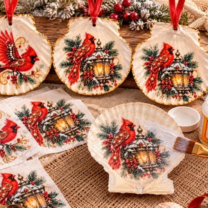 May include: Three scallop shell ornaments featuring red cardinal birds and lanterns, with matching decoupage paper squares, a paintbrush, and a jar of Mod Podge Matte adhesive. Each shell is topped with a red ribbon for hanging.