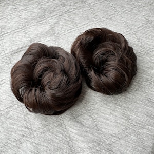 Puede incluir: Dos extensiones de moño de color marrón oscuro hechas de fibras sintéticas, dispuestas en un estilo circular, voluminoso y ondulado. Estas piezas de cabello tienen una textura natural con reflejos sutiles y una forma redondeada.