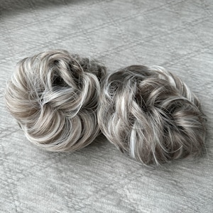 May include: Two circular hair bun extensions featuring a blend of light ash blonde, silver, and dark brown synthetic hair strands. The hair pieces are styled in a messy, voluminous circular shape with visible highlights and lowlights throughout.