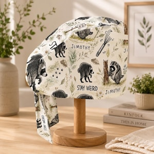 May include: A scrub cap featuring a cream-colored fabric with a repeating pattern of illustrated raccoons, green foliage, kitchen whisks, and the text "JIMOTHY" and "STAY WEIRD". The cap has a tie-back design and a structured, rounded crown shape.
