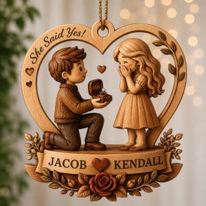 May include: A wooden heart-shaped ornament featuring a carved scene of a person kneeling to propose to another person. The text "She Said Yes!" is engraved at the top. A small box with "2026" and a ring is held out. A banner reads "JACOB" and "KENDALL" with a rose.