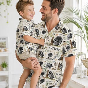 May include: Two short-sleeved button-down shirts featuring a repeating pattern of raccoons, trash cans, whisks, and trees on a light background. The shirts include the text "STAY WEIRD" and "JIMOTHY" printed in black among the various illustrations.