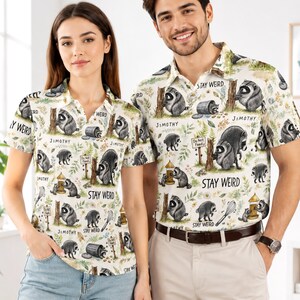May include: A short-sleeved polo shirt with a cream background featuring a repeating pattern of raccoons, bird feeders, trash cans, and whisks. The text "STAY WEIRD" and "JIMOTHY" is printed throughout the design, along with "Do Not Feed" signs.