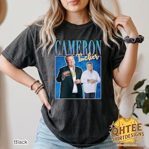 May include: A black short-sleeve t-shirt featuring a graphic print of the character Cameron Tucker from Modern Family. The design includes the text CAMERON TUCKER in blue and yellow, with two images of the character set against a blue background.