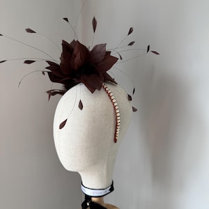 May include: A dark brown fascinator featuring a large, layered flower and delicate feather detailing. A sparkling rhinestone band curves across the headpiece. The headpiece is displayed on a mannequin head with a black ribbon.