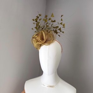 May include: A gold-coloured fascinator with a floral design and feather accents. The headpiece is displayed on a white mannequin head. The headband is a thin, light brown colour. Suitable for special occasions.