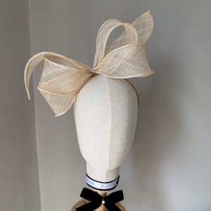 May include: A beige headpiece featuring a large, woven bow. The bow is positioned on a mannequin head. A black velvet bow and a ribbon with text are around the mannequin's neck. The headpiece is suitable for a special event.