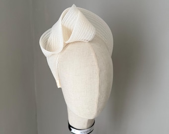 May include: An ivory-coloured hat with a net overlay, displayed on a mannequin head. The hat has a looped design on top. The mannequin head is on a wooden stand with a black metal base. The hat is a formal accessory, suitable for special occasions.