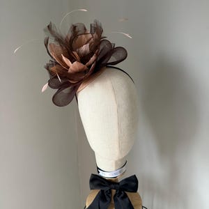 May include: A brown and tan floral fascinator with feathers, set on a black headband. The headpiece is displayed on a mannequin head with a black bow and a white ribbon around the neck.