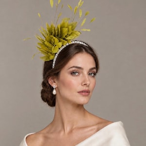 May include: A headband featuring a cluster of olive green feathers and thin, upright stems. The band is encrusted with small, clear, sparkling crystals that line the entire top edge, providing a reflective and detailed finish to the accessory.