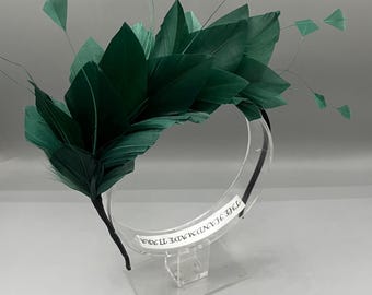 May include: A dark green feather and leaf headband. The headband features large, dark green feathers and leaves, with smaller square-shaped accents. The headband is on a clear display stand. The text "THE HANDMADE TIARA" is visible on the headband.