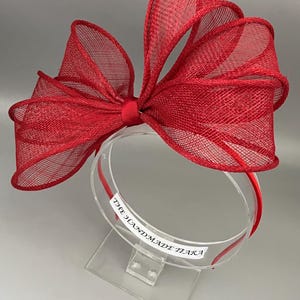 May include: A red mesh headband with a large bow. The headband is on a clear display stand with the text "The Handmade Tiara" on the stand.