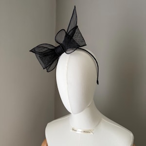 May include: A black headband with a large, structured bow made from a sheer, textured material. The bow is the main feature, with a layered design and a prominent knot. The headband is displayed on a mannequin head against a neutral backdrop.