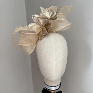 May include: A beige headpiece with a floral design. The headpiece features a large bow and is adorned with metallic gold and silver flower accents. The headpiece is displayed on a mannequin head with a black ribbon.