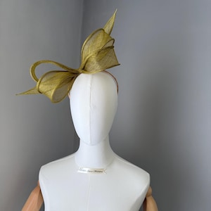 May include: A gold headpiece featuring a large bow design, mounted on a thin headband. The headpiece is displayed on a white mannequin against a grey background. The bow has a textured, woven appearance.