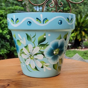May include: A light blue ceramic planter with a white and green painted floral design. The planter features white daisies and blue flowers with green leaves.