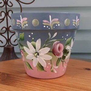 May include: A hand-painted ceramic flower pot with a blue and pink ombre design. The pot features a floral pattern with white flowers, pink roses, and green leaves.