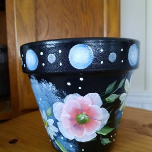 May include: A black terracotta flower pot with white polka dots and blue circles around the rim. The pot is decorated with hand-painted pink and white flowers.