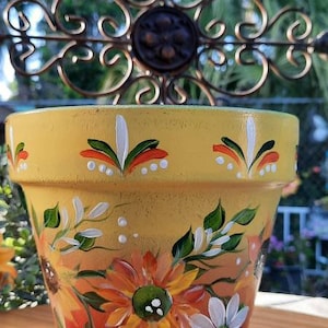 May include: Hand-painted terracotta flower pot with a yellow base, red and orange flowers, and white accents. The pot is decorated with green leaves and white dots.