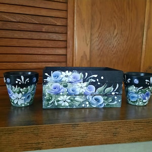 May include: Three-piece set of hand-painted floral decor. The set includes two small black pots and a rectangular box. Each piece is decorated with blue roses, white daisies, and green leaves. The background is a dark wood.