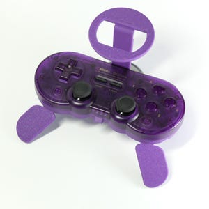 May include: A translucent purple 8BitDo SN30 Pro wireless game controller featuring a directional pad, two black analog sticks, four action buttons labeled A, B, X, and Y, and a purple plastic smartphone mount attached to the top center.