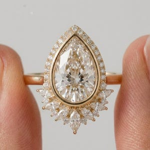 Puede incluir: Un anillo de oro con una gran piedra central en forma de pera rodeada por un halo de pequeños diamantes redondos. La base del engaste está acentuada con un grupo de diamantes de talla marquesa. La palabra CORVEN está grabada en el anillo.