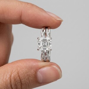 Puede incluir: Un anillo de tono plateado con una piedra central de corte cuadrado y un patrón geométrico concéntrico. La banda está compuesta por dos hilos entrelazados, cada uno incrustado con pequeñas piedras transparentes en pavé hacia el centro.
