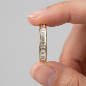Puede incluir: Un anillo de banda dorada con una fila continua de piedras transparentes de corte baguette engastadas en canal. El anillo tiene un acabado metálico pulido y un ancho uniforme, mostrando una serie de piedras rectangulares dispuestas verticalmente.