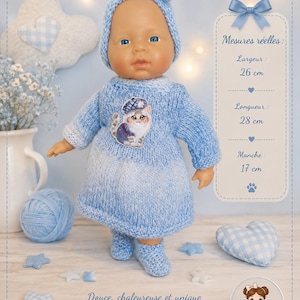 Op de afbeelding: Een pop draagt een lichtblauw gebreid jurkje, bijpassende muts en slofjes. De jurk heeft een kattenapplicatie. Een label toont de afmetingen: breedte 26 cm, lengte 28 cm en mouw 17 cm. De pop heeft blauwe ogen en een lichte huidskleur.
