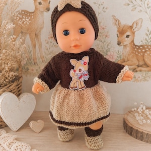 May include: A doll wearing a dark brown knitted hat with a small bow, a dark brown long-sleeved sweater featuring a fawn patch on the chest, and a light beige knitted skirt. The doll also wears matching light beige knitted booties on its feet.