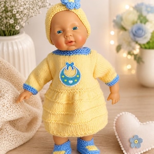 May include: A baby doll wearing a yellow knitted dress with a blue bib applique, a matching yellow hat with a blue flower, and blue and yellow knitted booties. The doll has light skin, blue eyes, and a neutral expression, featuring a textured knit.