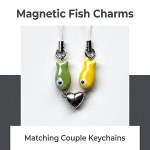May include: Magnetic Fish Charms Matching Couple Keychains featuring one green and one yellow fish-shaped charm, each with a white eye, attached to a silver-toned metal heart that connects magnetically at the base with clear cord loops.