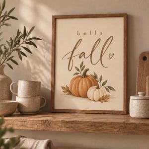 Può includere: Una stampa artistica rettangolare da parete in una cornice di legno marrone con il testo hello fall in caratteri corsivi e stampatelli marrone scuro, accompagnata da un'illustrazione di una grande zucca arancione e una zucca bianca più piccola.