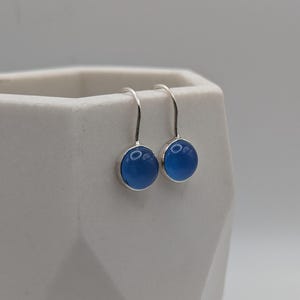 Könnte beinhalten: Ein Paar silberne Ohrhänger mit runden, hellblauen Cabochon-Steinen. Die Ohrringe sind in einer einfachen Lünettenfassung gehalten und hängen an einem gebogenen Silberdraht. Die Ohrringe werden vor einem weißen Hintergrund präsentiert.