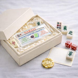 Pode incluir: Uma caixa de presente cor de creme contendo um cartão de Mahjong de acrílico transparente com o nome SARAH impresso em vermelho, juntamente com várias peças de Mahjong, dois dados vermelhos e uma ficha circular dourada com centro branco.
