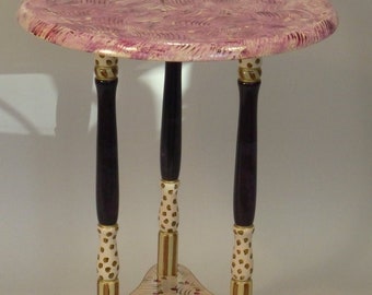 May include: A small, round, purple and white painted table with three legs. The legs are painted black with gold and white accents. The table top has a swirling pattern.