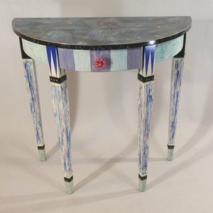 May include: A half-round table with a single drawer and three legs. The table is painted in shades of blue, white, and black with a decorative pattern. The drawer has a pink and red flower-shaped knob.