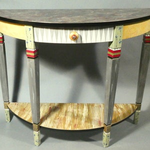May include: A half-round table with a gray top, a drawer with a geometric pattern, and three legs with decorative details. The legs are painted gray with red and gold accents. The table has a lower shelf.