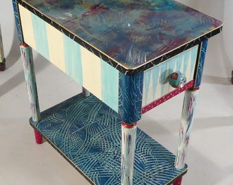 May include: A small, hand-painted side table with a single drawer and a lower shelf. The table is painted in a blue, white, and gold color scheme with a whimsical, abstract design. The legs are painted in a white and red striped pattern.