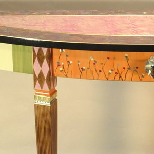 May include: A half-round table with a painted wood top and a single drawer. The table is painted in a variety of colors, including green, white, pink, brown, and orange. The drawer has a floral design and a decorative knob.