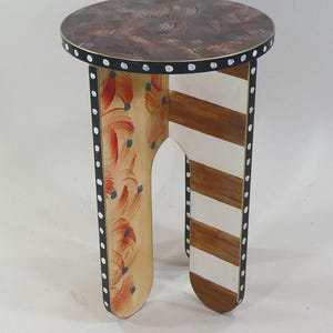 May include: A small round side table with a dark brown textured top. The edges are black with white dots. One leg features orange brushstrokes with blue dots on a light wood background, while the other leg has horizontal brown and white stripes.