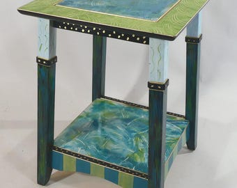 May include: A small, square end table with a green top and blue and green painted legs. The table has a lower shelf and is decorated with black and gold accents.