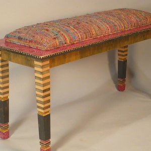 May include: A wooden bench with a colorful woven fabric cushion. The bench has black and gold striped legs with pink accents.